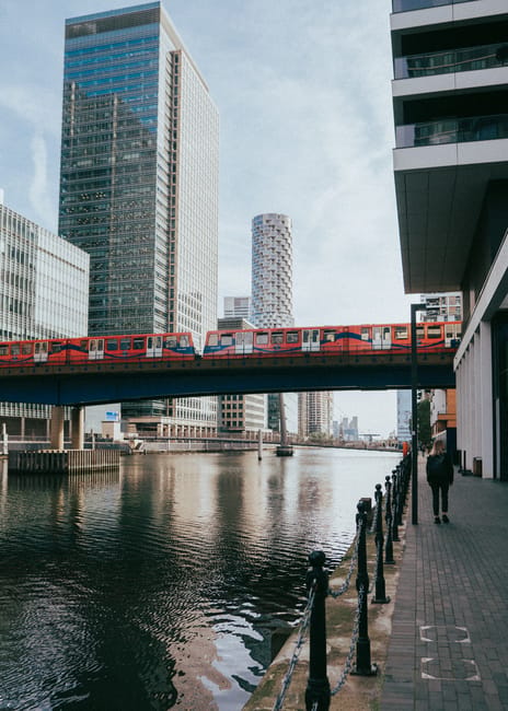 A view of a modern urban canal scene in Canary Wharf with high-rise office buildings featuring glass and steel facades. A red train crosses a bridge above the waterway, which has a reflective, calm surface. Along the canal, a paved walkway with black metal bollards and a person walking is visible on the right, with the buildings casting structured shadows on the pavement. The scene illustrates a clean, well-maintained city environment. Cleaners E8 specialise in surface cleaning and deep cleaning solutions suited for commercial premises like office corridors and public walkways, ensuring hygiene and maintaining the appearance of busy urban areas such as Canary Wharf.