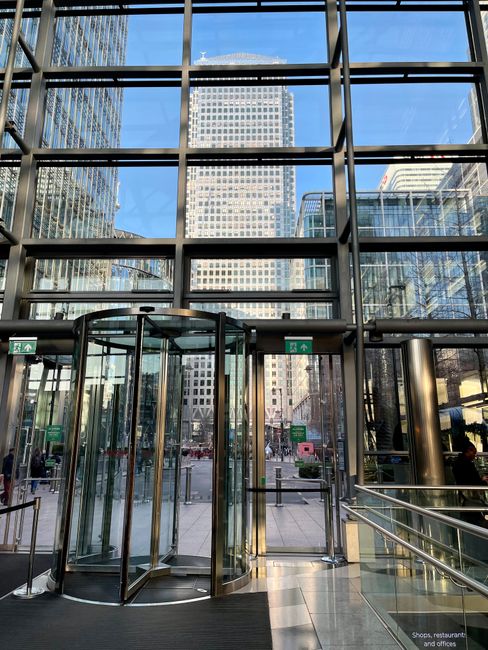 A wide view of the Canary Wharf area showcasing modern high-rise buildings with glass and steel facades lining a waterway. The scene features a mix of architectural styles, including a cylindrical tower with a distinctive patterned exterior. The buildings are illuminated by natural daylight, casting reflections on the calm water surface. In the foreground, the waterway is bordered by a walkway and some visible infrastructure elements. This urban landscape exemplifies contemporary city architecture, relevant to residential and commercial cleaning services offered by Cleaners E8 for high-rise surfaces, windows, and exteriors, emphasizing surface cleaning and sanitisation in modern office environments.
