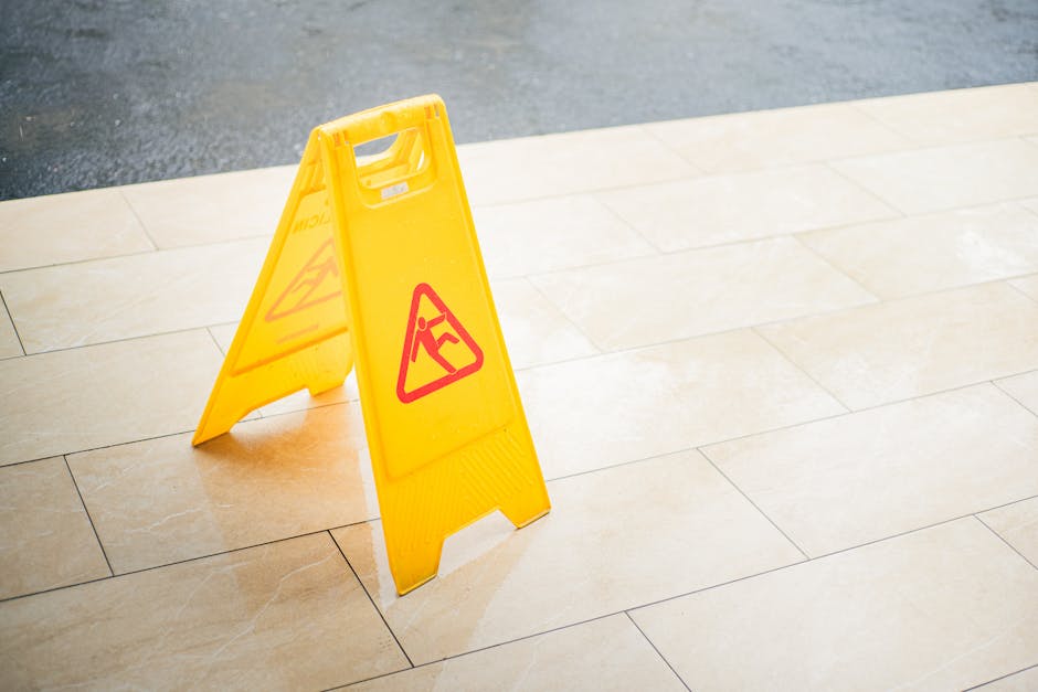 A bright yellow wet floor warning sign positioned on a tiled hallway floor, indicating a slip hazard. The tiles are smooth, beige, and show a clean surface with a polished appearance. The sign is plastic with a red and black warning symbol, and it stands upright on the floor, which is part of a corridor or entrance area. The lighting highlights the glossy finish of the tiles, and there are no visible dust or stains, suggesting recent cleaning or maintenance. This scene demonstrates surface cleaning and safety measures, aligning with hygienic and upkeep procedures that Cleaners E8 provides for residential or commercial spaces, particularly in areas like foyers or corridors.