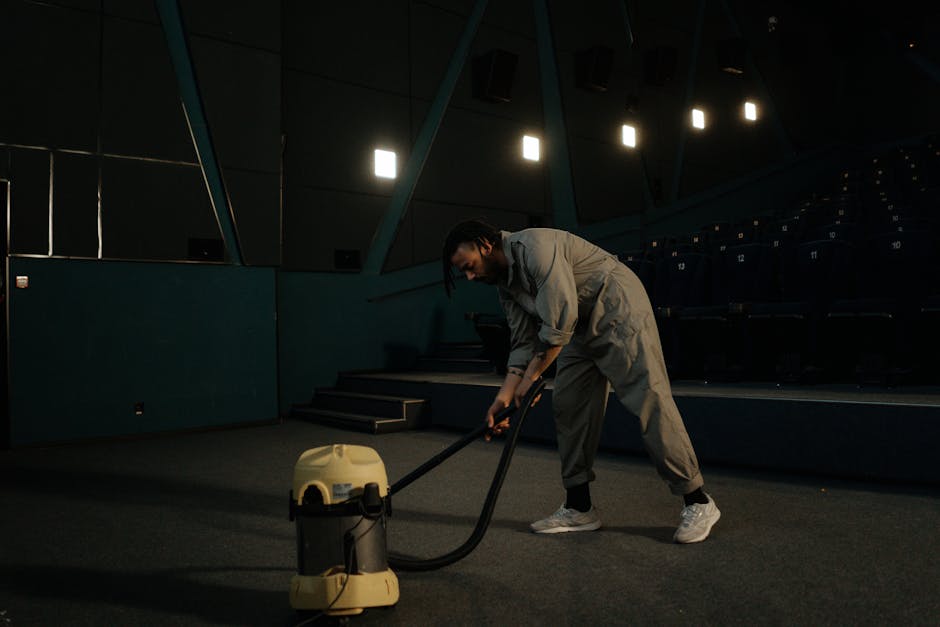 A professional cleaner from Cleaners E8 is performing surface cleaning or deep cleaning in an auditorium or conference room. The individual, dressed in a grey jumpsuit, is using a floor vacuum or carpet cleaner with a long hose attachment on a dark carpeted surface. The room features tiered seating with navy blue chairs arranged in rows, illuminated by overhead lighting arranged in a linear pattern, with reflective wall panels contributing to the modern design. The cleanliness of the carpet is emphasized, with no visible dust or debris, indicating a thorough sanitisation process. The setting is dimly lit, focusing on the cleaning activity, highlighting the importance of hygiene and maintenance for commercial spaces. The image underscores the expertise of Cleaners E8 in professional deep cleaning and surface sanitation services.