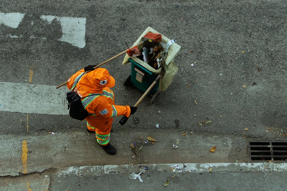 A sanitation worker dressed in bright orange high-visibility clothing, including a jacket and trousers with reflective stripes, is cleaning a city street using a long-handled broom. The worker is push