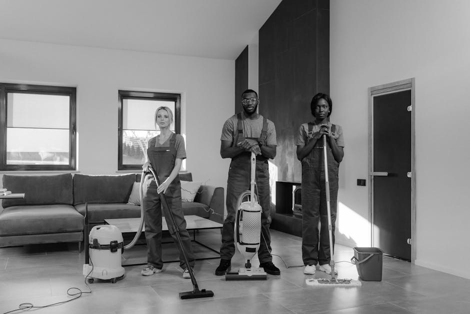 Three professional cleaners from Cleaners E8 are performing surface cleaning and deep cleaning tasks in a modern living room with natural light coming through two rectangular windows. The room features a large, L-shaped sofa with a fabric cover, a coffee table, and a built-in wood panel against the wall. The workers are equipped with vacuum cleaners and mops, wearing protective aprons, and are focused on their cleaning tasks. The floor appears to be laminate or hardwood, visibly clean and polished, reflecting the light. The surrounding surfaces, including the sofa arms and the floor, look dust-free and well-maintained, indicating thorough sanitisation and hygiene. The scene emphasizes domestic cleaning and surface care, showcasing meticulous cleaning processes in a residential environment, aligned with the services provided by Cleaners E8 in the Docklands E8 area.