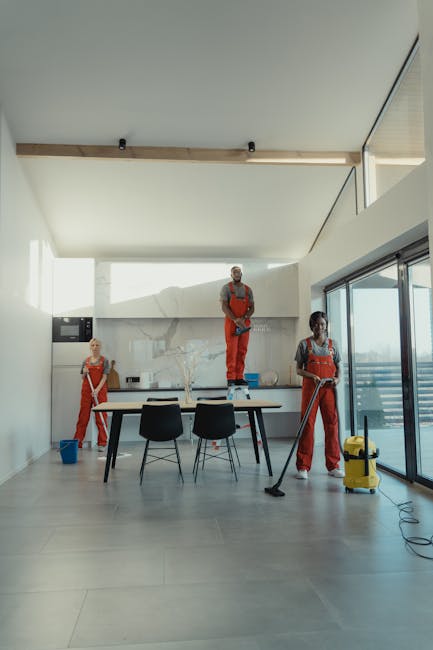A modern dining area with a large rectangular table surrounded by four black chairs on a light grey tiled floor. In the background, three professional cleaners dressed in red overalls and white shirts are performing deep cleaning tasks. One cleaner stands on a chair, cleaning the upper part of a white wall, while another stands on the floor with a vacuum cleaner, and the third is wiping the window area near sliding glass doors that lead to a balcony. The room features a white ceiling with wooden beam accents and ample natural light coming through the large glass doors, highlighting the clean surfaces and organized space, with minimal decor including a decorative bowl on the table. Cleaners E8 provide thorough domestic cleaning, ensuring surfaces such as floors, windows, and walls are hygienic and polished in line with top-rated end of tenancy cleaning standards.