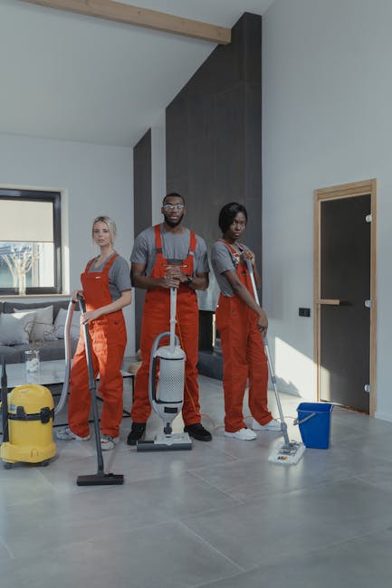 Three professional cleaners from Cleaners E8 are performing surface cleaning and deep cleaning tasks in a modern living room with natural light coming through two rectangular windows. The room features a large, L-shaped sofa with a fabric cover, a coffee table, and a built-in wood panel against the wall. The workers are equipped with vacuum cleaners and mops, wearing protective aprons, and are focused on their cleaning tasks. The floor appears to be laminate or hardwood, visibly clean and polished, reflecting the light. The surrounding surfaces, including the sofa arms and the floor, look dust-free and well-maintained, indicating thorough sanitisation and hygiene. The scene emphasizes domestic cleaning and surface care, showcasing meticulous cleaning processes in a residential environment, aligned with the services provided by Cleaners E8 in the Docklands E8 area.