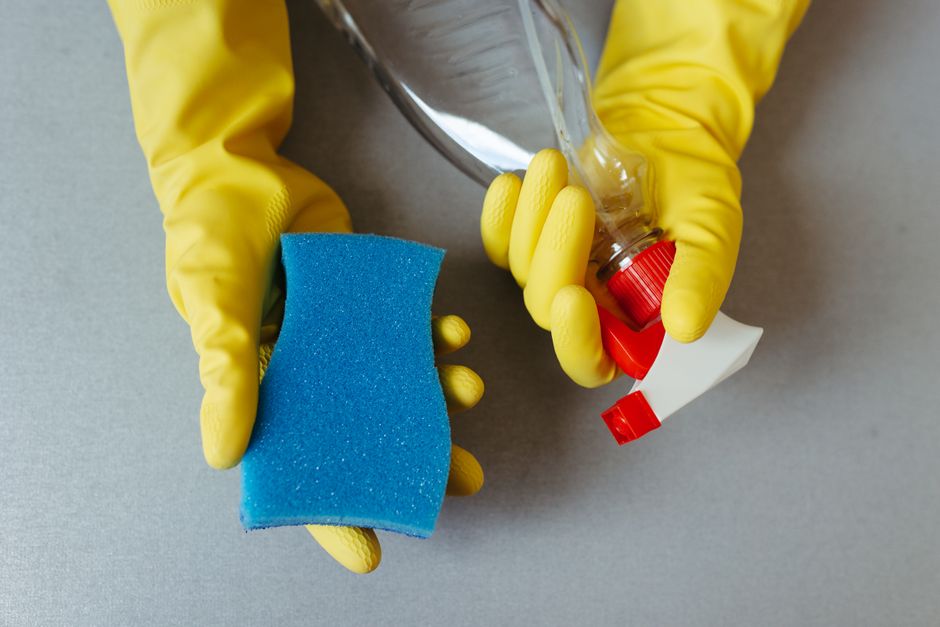 A pair of gloved hands holding a blue cleaning sponge in one hand and a spray bottle with a red cap and a white spray nozzle in the other. The sponge appears slightly wet and is used for surface cleaning, with the spray bottle likely containing a cleaning solution. The background is a plain, neutral surface, emphasizing the cleaning tools. This image illustrates a typical domestic cleaning process involving surface sanitation and deep cleaning to maintain hygiene, aligning with the focus on cleaning principles discussed on the page 'Tower Hamlets cleaning rules landlords need to know E8, E8' by Cleaners E8.