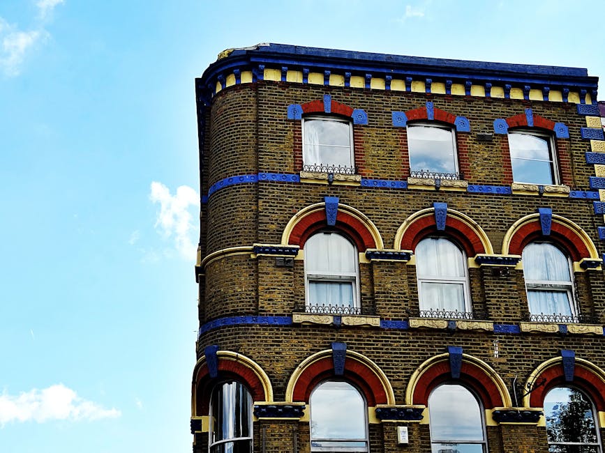 Close-up view of the upper floors of a historic brick building in Tower Hamlets, E8, with multi-pane windows framed by red and beige arches, decorative blue and yellow accents along the cornice and window trims, and a clear blue sky with a few white clouds in the background. The building's exterior appears clean and well-maintained, with glass windows reflecting the sky, indicating proper surface cleaning and preservation, as part of ongoing maintenance in line with Tower Hamlets cleaning rules landlords need to know. The image showcases the architectural details and the pristine condition of the facade, emphasizing the importance of thorough surface cleaning and building upkeep by companies such as Cleaners E8.